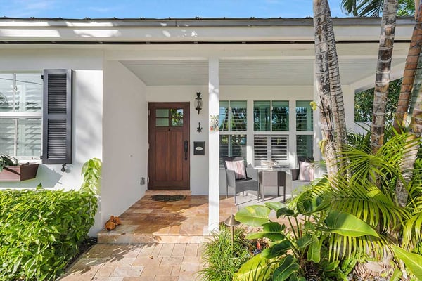 Modern white house exterior with covered porch, dark wood door, palm trees, and tropical plants in landscaping