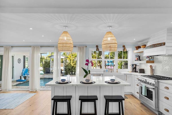 Bright kitchen with white cabinetry, woven pendant lights, island with black stools, and view to pool area through glass doors